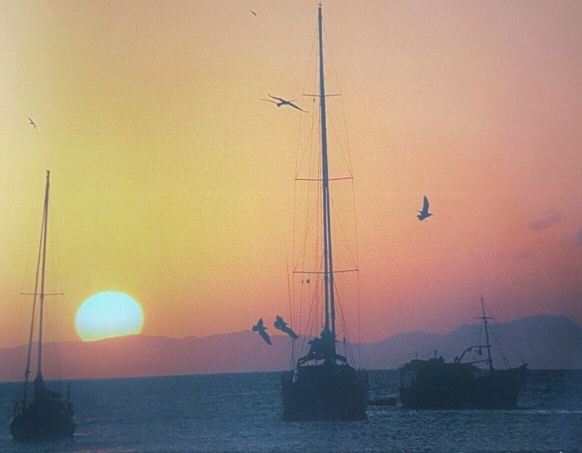 A sunset over a calm sea with sailboats and birds flying near the horizon.