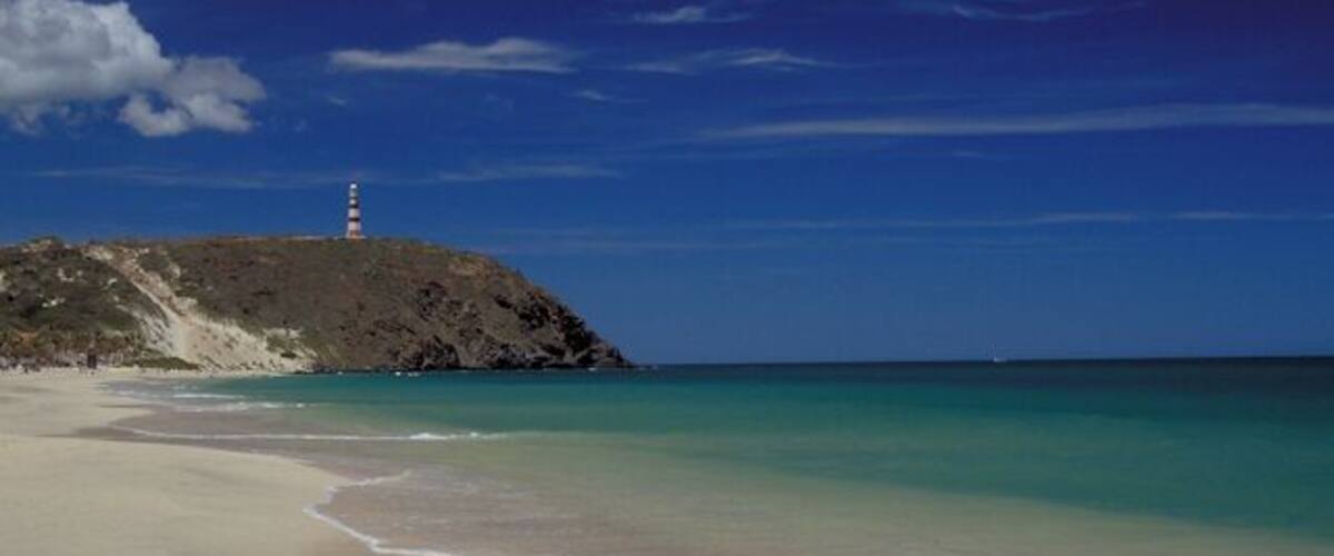 A beach with clear turquoise water, a sandy shore, and a lighthouse on a rocky hill.