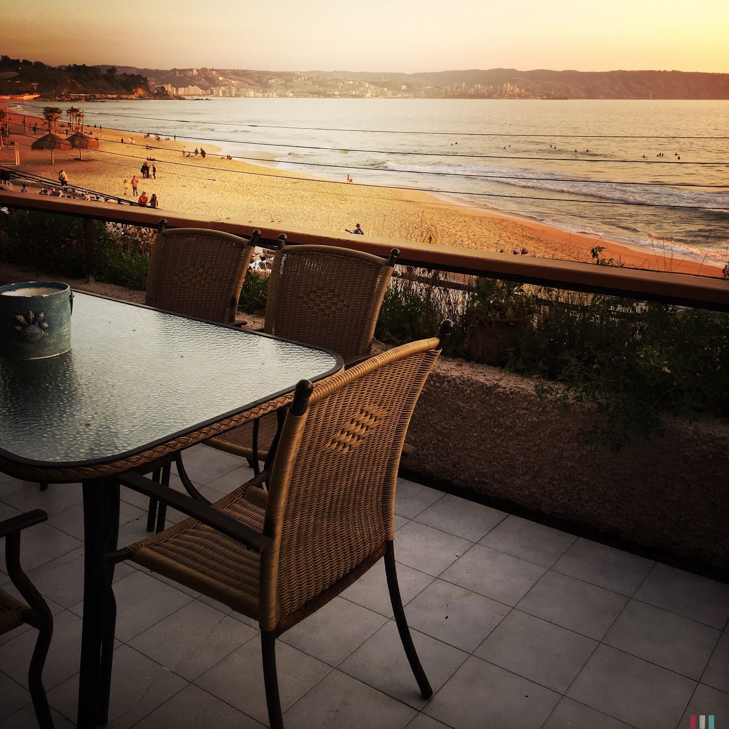 A balcony with wicker chairs overlooking a beach with people and a cityscape in the background.