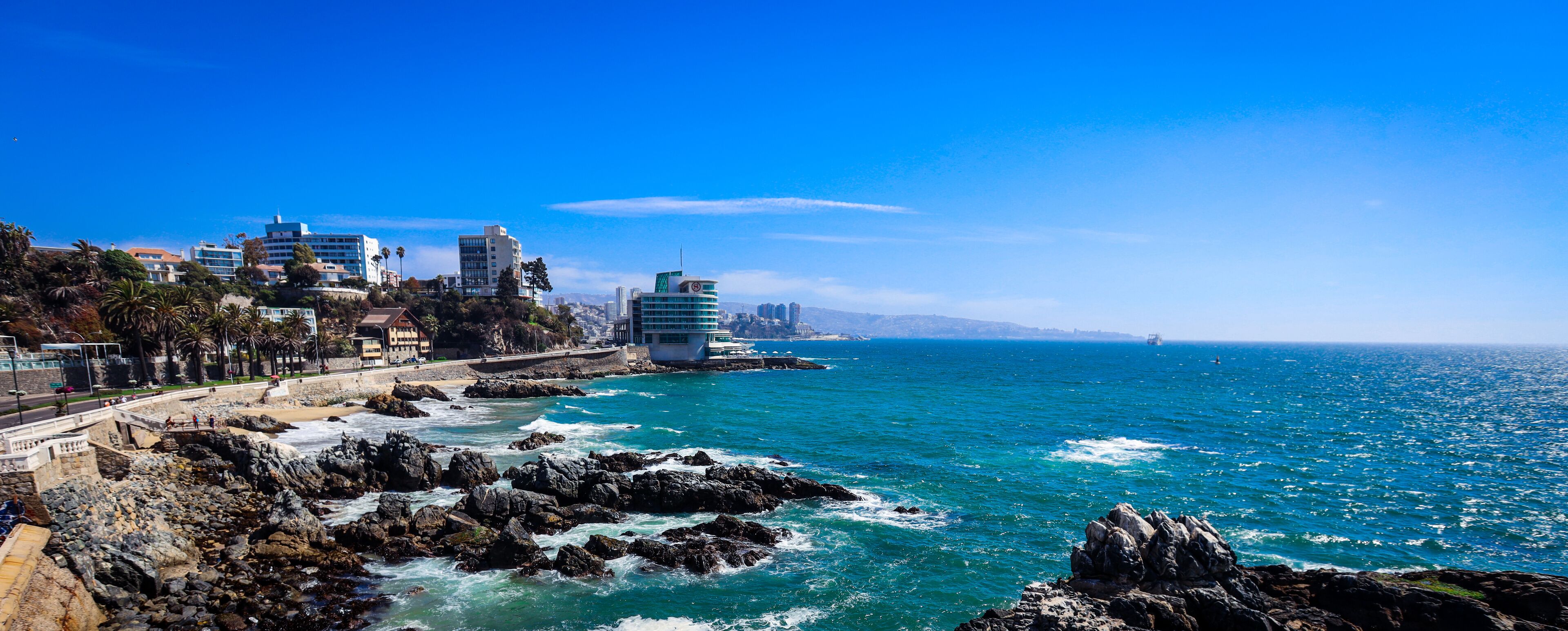A coastal cityscape with rocky shores, buildings, and a clear blue sky.
