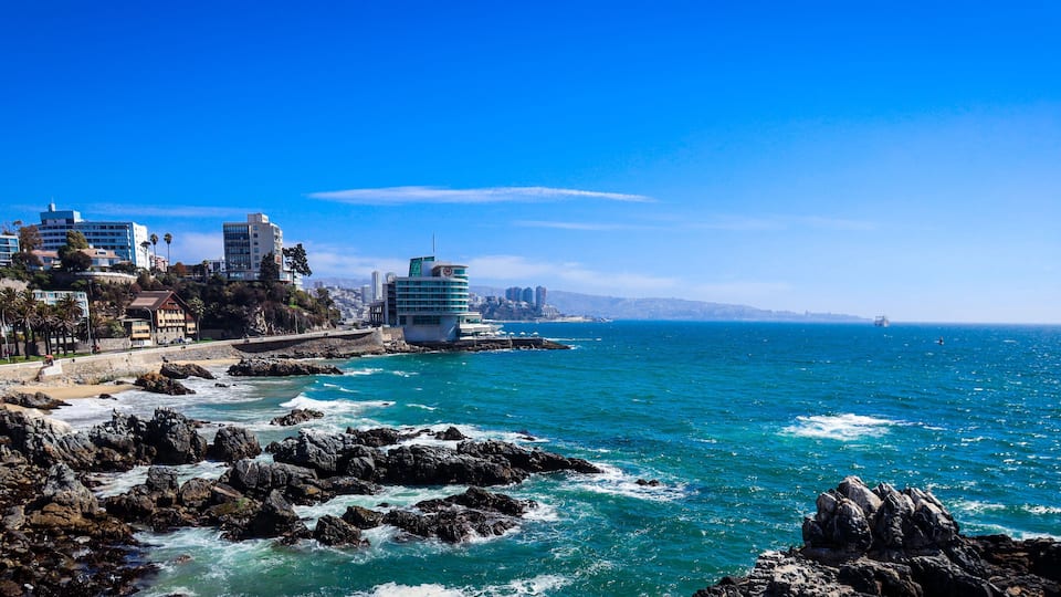 A coastal cityscape with rocky shores, buildings, and a clear blue sky.