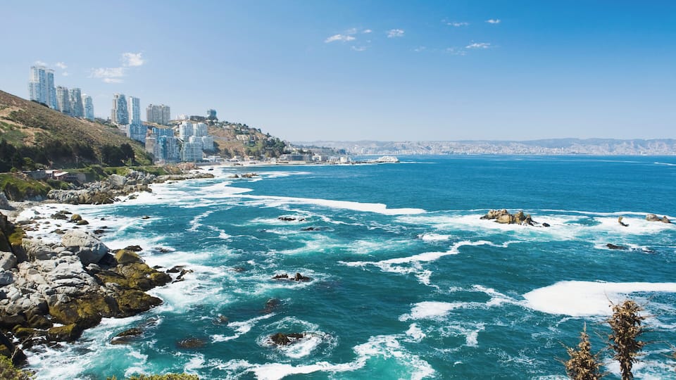 A coastal city with a rocky shoreline, clear blue sea, and high-rise buildings on a hillside.