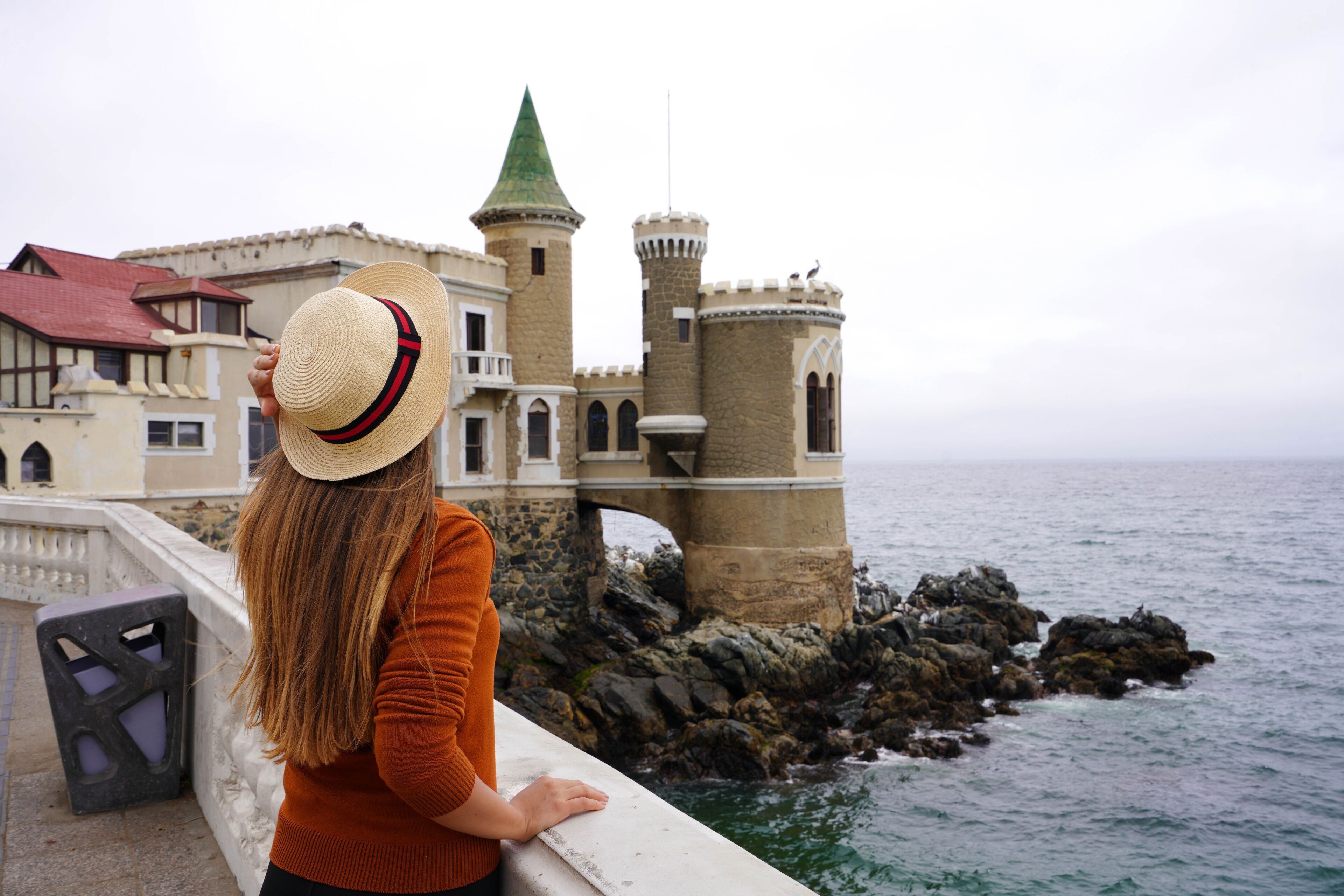 Un castillo costero con una aguja verde, un puente de piedra y un litoral rocoso.