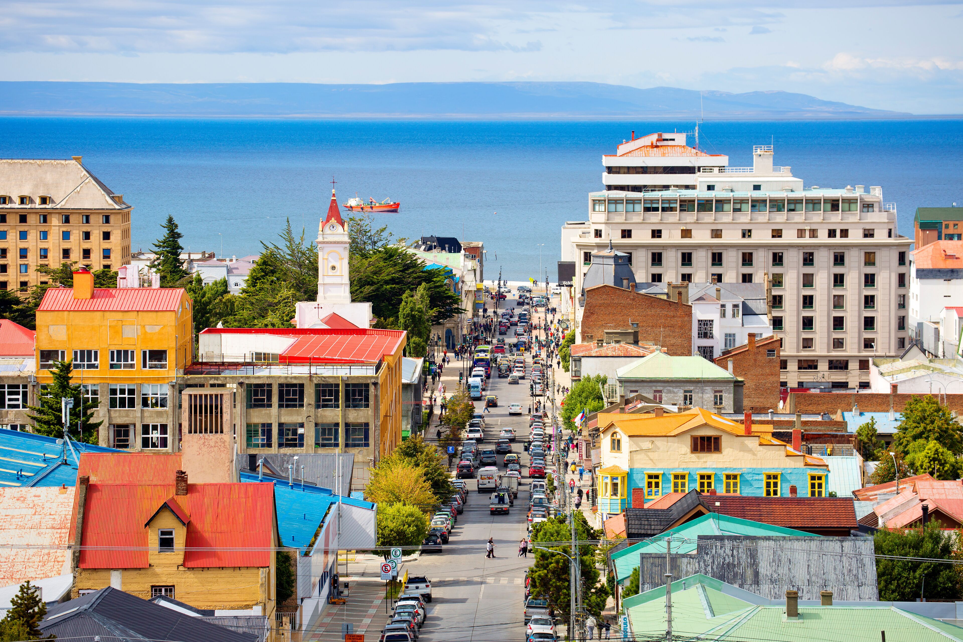 Paisagem costeira com edifícios coloridos, rua movimentada e um navio ao longe.