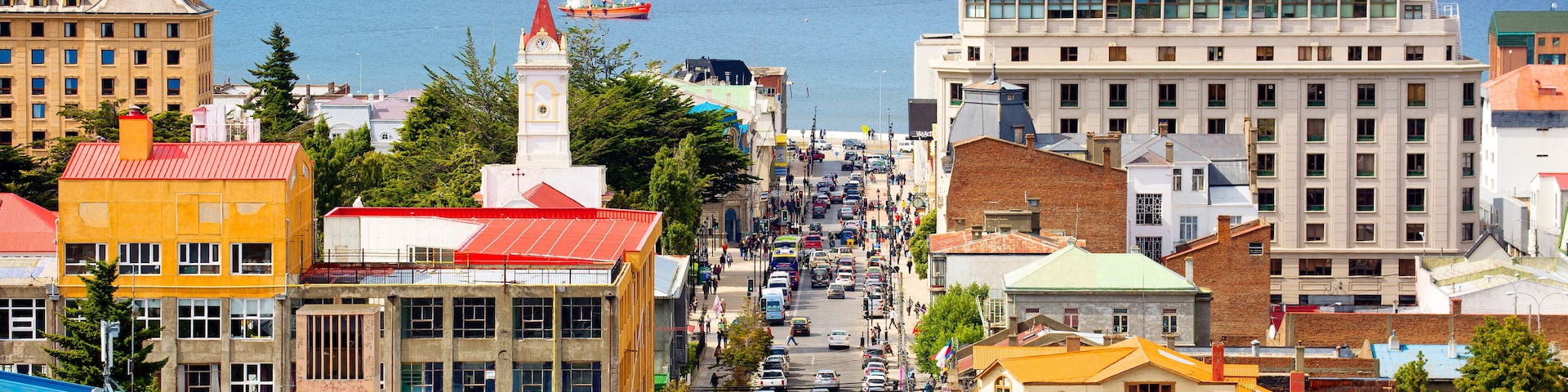 Paisagem costeira com edifícios coloridos, rua movimentada e um navio ao longe.
