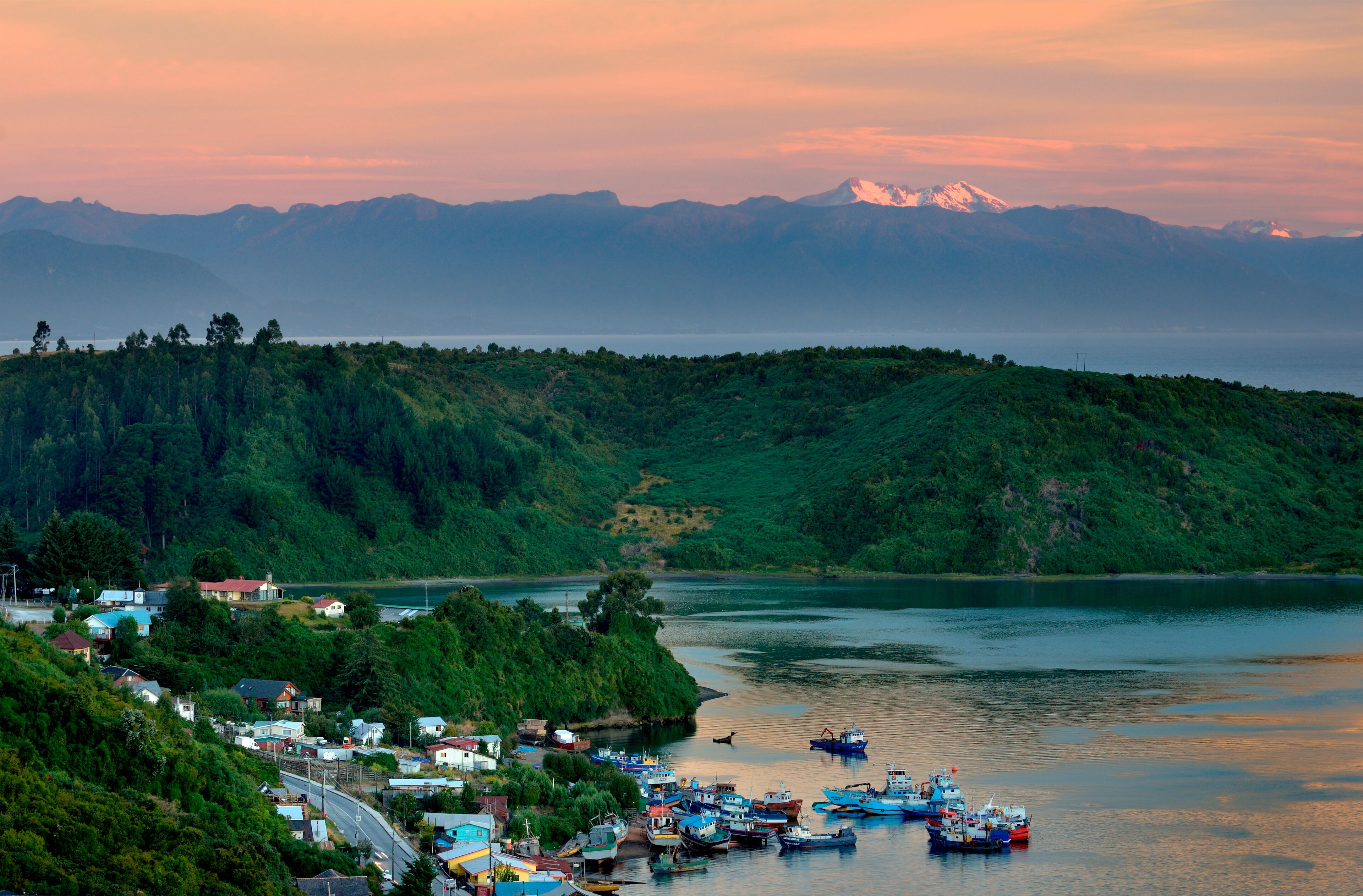 A coastal village with boats docked along the shore, surrounded by lush green hills and mountains in the background.