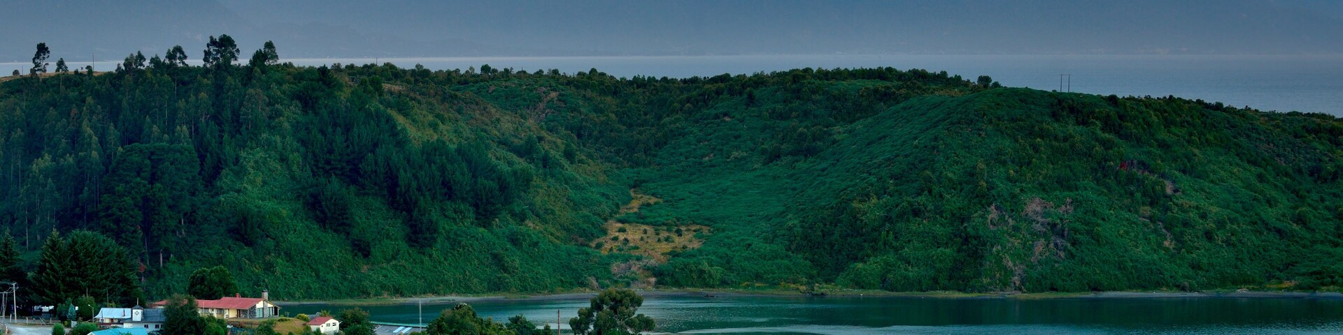 A coastal village with boats docked along the shore, surrounded by lush green hills and mountains in the background.