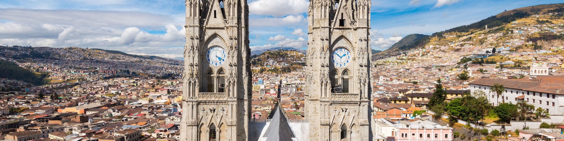 A cathedral with two towers and a clock, surrounded by a cityscape and hills.