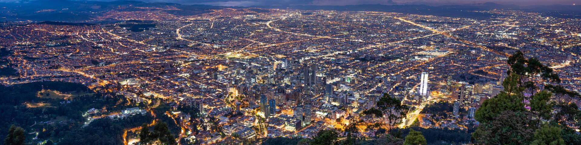 A panoramic view of a city at night with illuminated streets and buildings, surrounded by mountains and a cloudy sky.