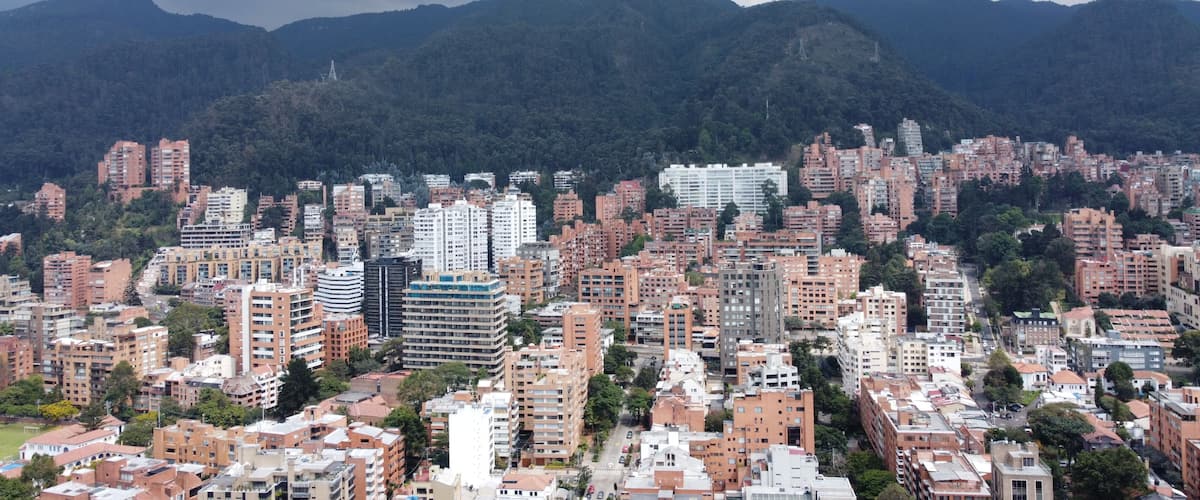 Un paisaje urbano con edificios residenciales densos, un rascacielos blanco destacado y montañas al fondo.
