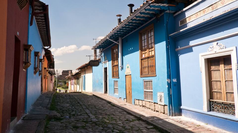 Una calle empedrada flanqueada por edificios de colores, incluyendo un edificio azul con ventana blanca y uno rojo con puerta marrón.