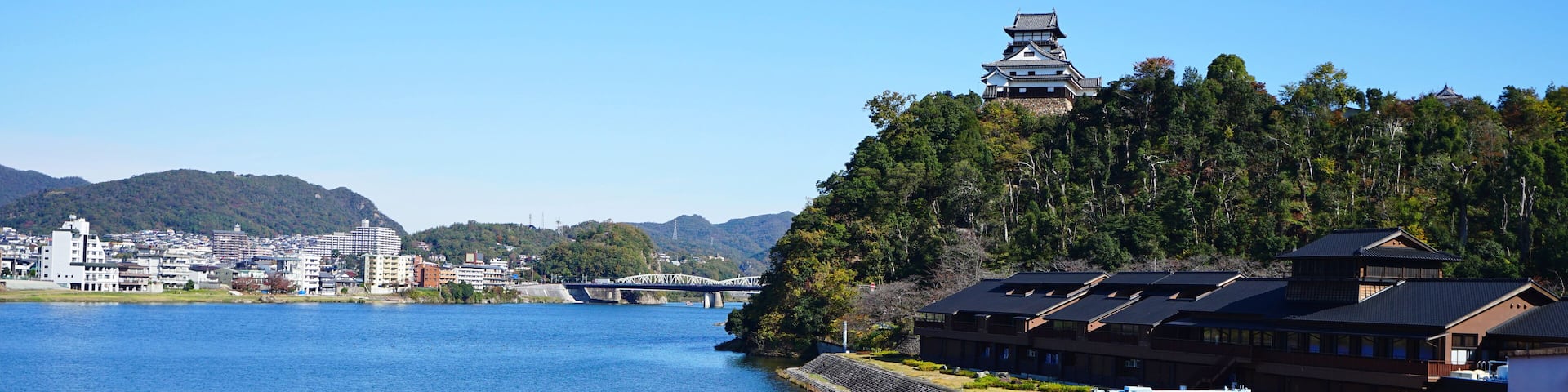 A traditional Japanese pagoda on a hill overlooking a river, with modern buildings and a bridge in the background.