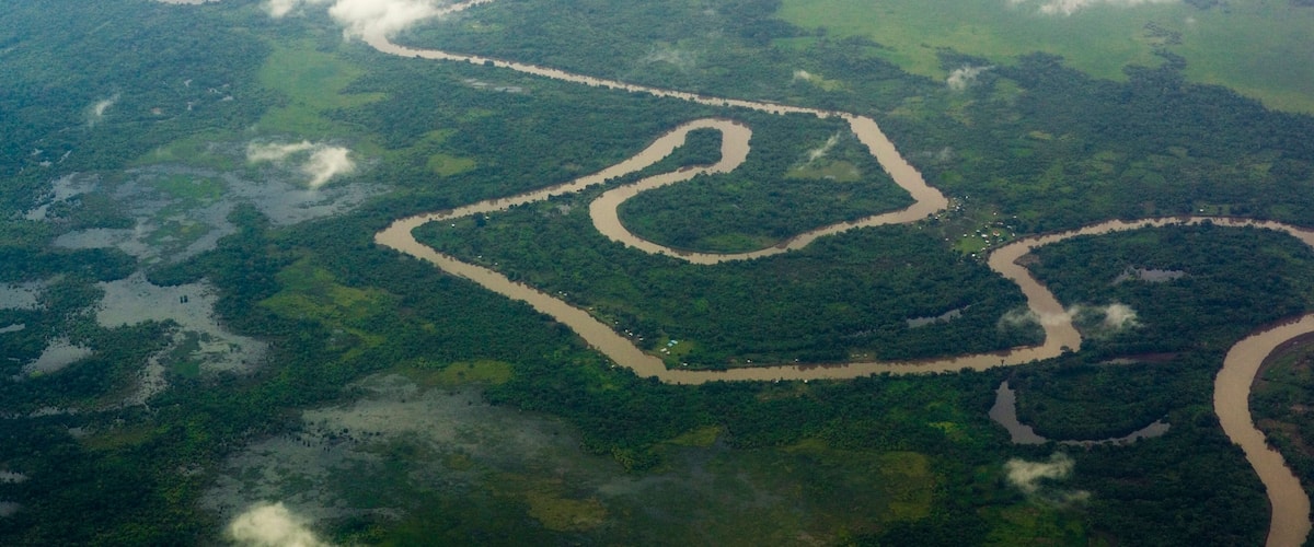 An aerial view of a river winding through a dense forest with clearings and a winding road.