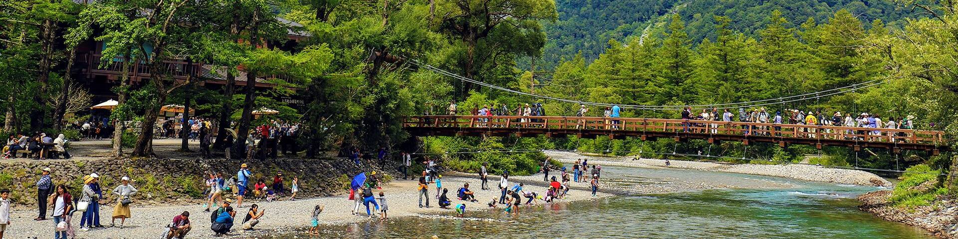 A wooden bridge over a river with people walking and a mountainous landscape in the background.