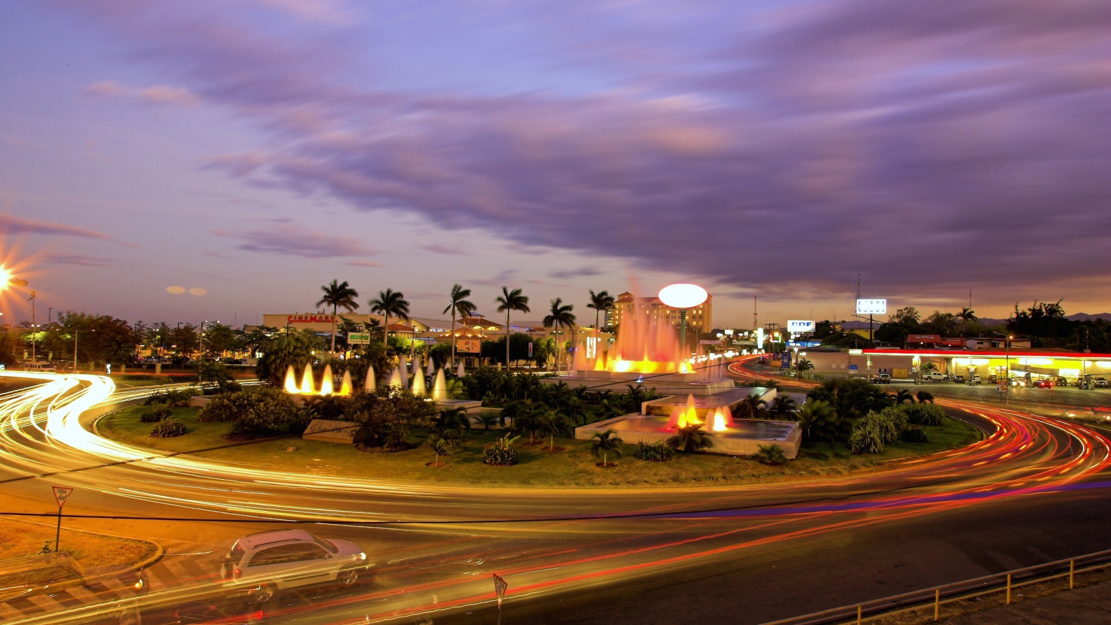 A cityscape at dusk with a prominent fountain, palm trees, and a busy road with light trails.