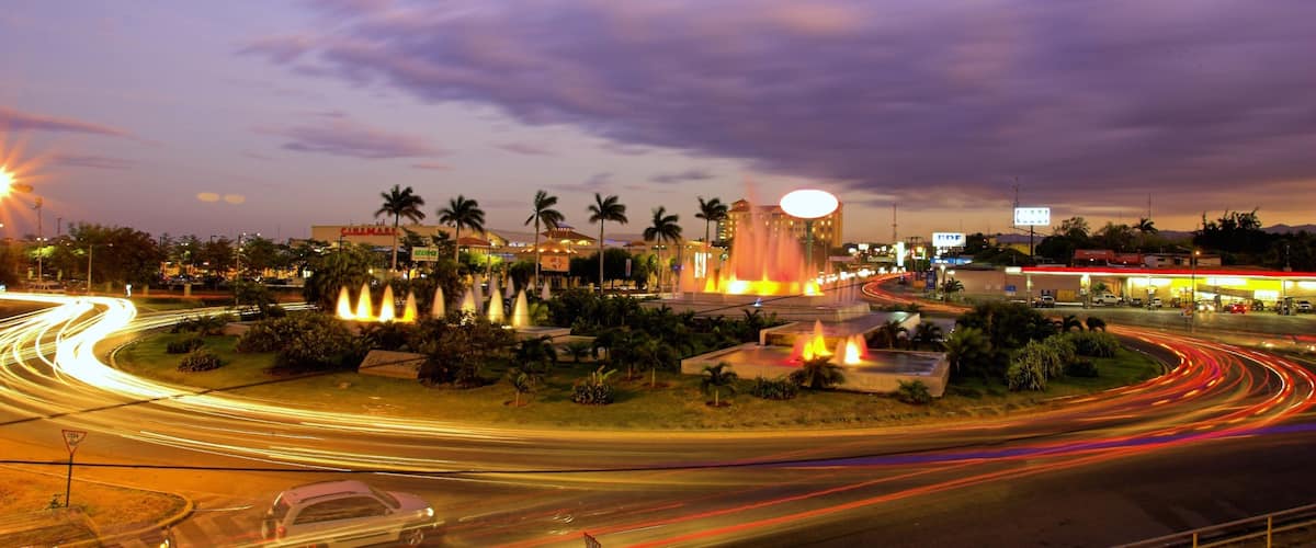 A cityscape at dusk with a prominent fountain, palm trees, and a busy road with light trails.