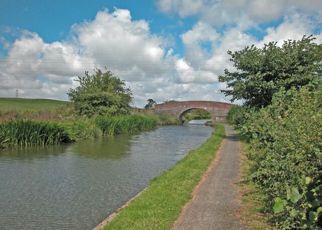 A narrow canal with a brick bridge, surrounded by greenery and a clear sky.