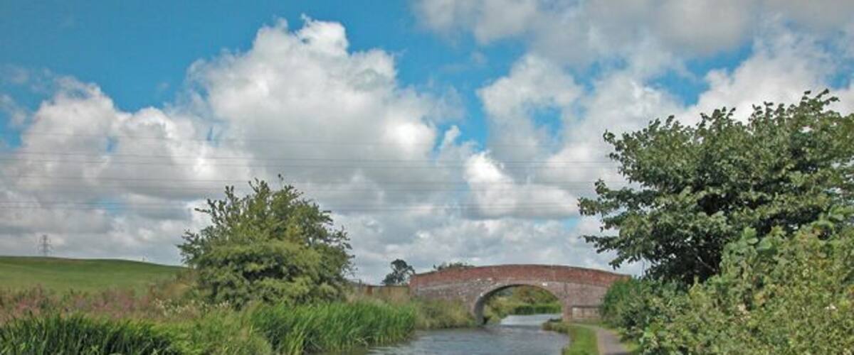 A narrow canal with a brick bridge, surrounded by greenery and a clear sky.