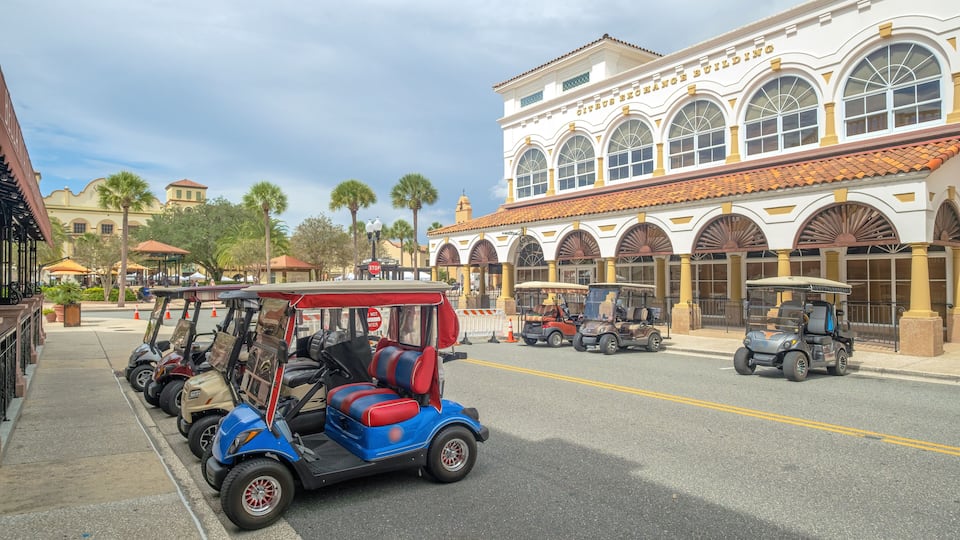 A street with golf carts parked on the sidewalk, a building with arched windows, and palm trees in the background.