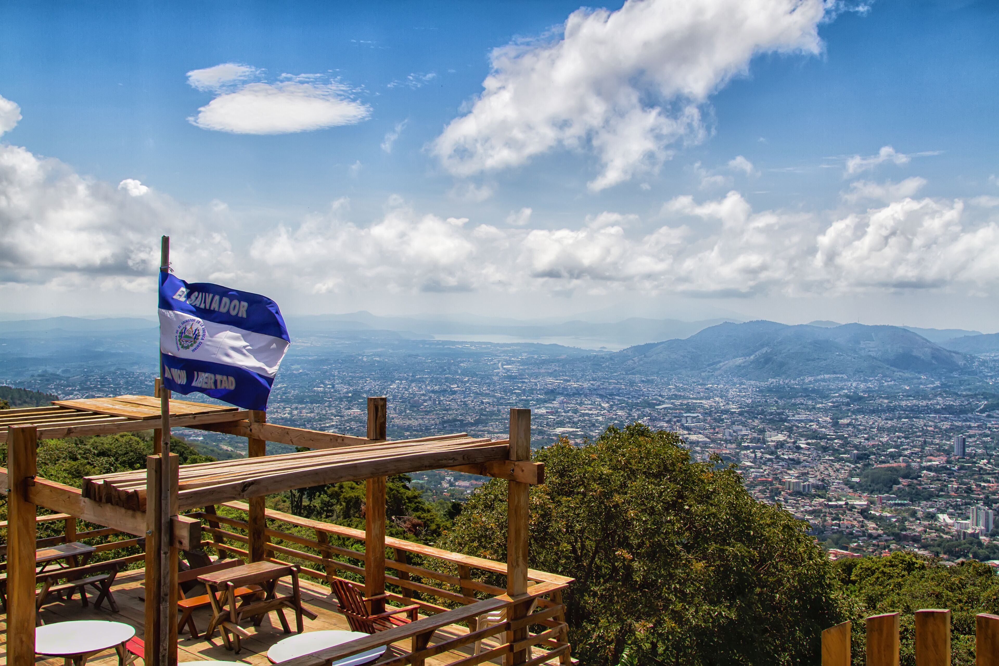 A wooden deck with benches overlooking a cityscape and mountains. A flag of El Salvador is visible.
