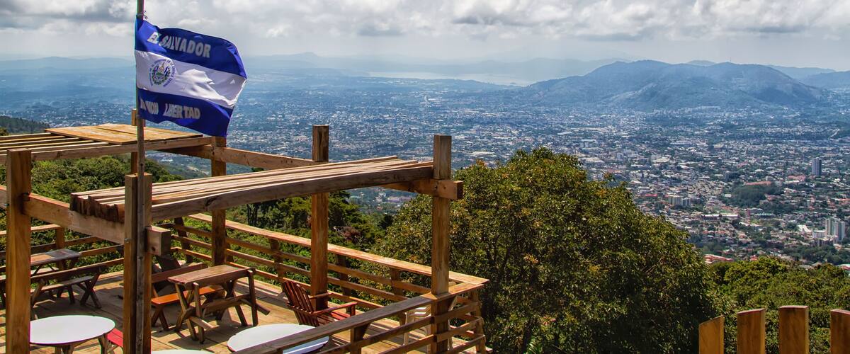A wooden deck with benches overlooking a cityscape and mountains. A flag of El Salvador is visible.