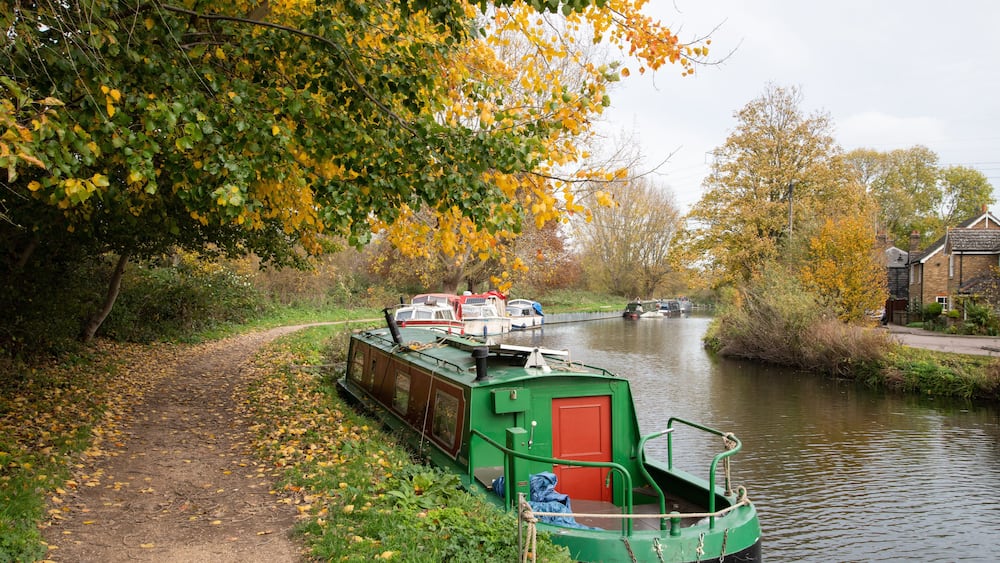 A canal boat cruising the River Lea Navigation between Enfield Lock and Waltham Cross, surrounded by trees in warm autumn colours.