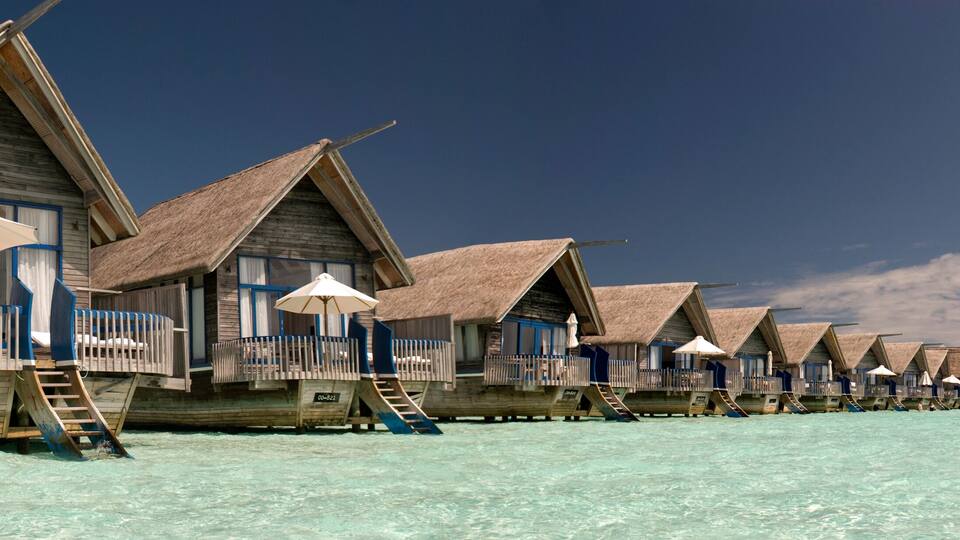 Overwater bungalows with thatched roofs and wooden railings on a clear turquoise sea.