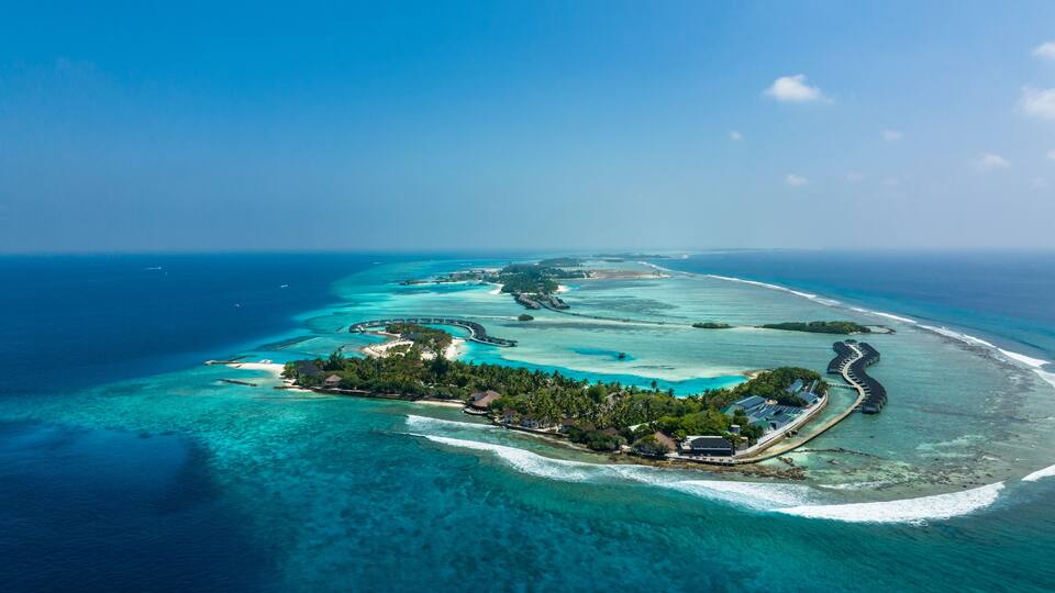 An aerial view of a tropical island with clear turquoise waters, a crescent-shaped resort, and a long pier extending into the sea.