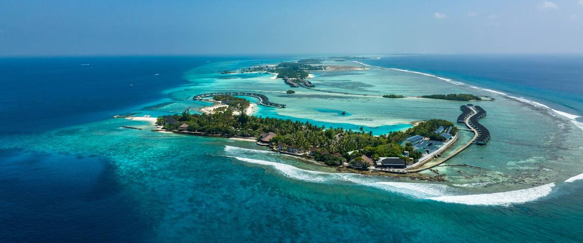 An aerial view of a tropical island with clear turquoise waters, a crescent-shaped resort, and a long pier extending into the sea.
