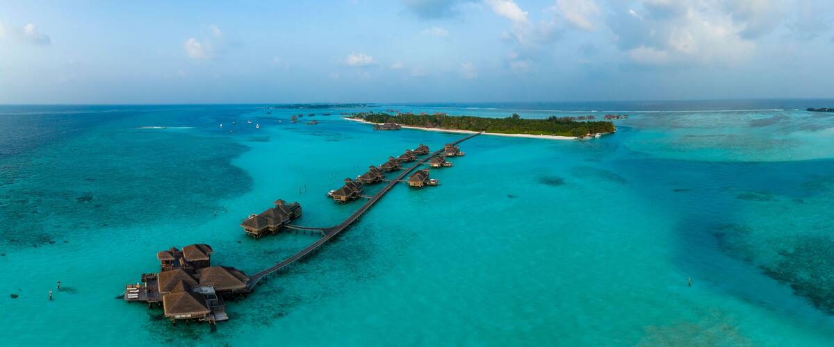 Vista aérea de un resort tropical con bungalows sobre el agua, unidos por una pasarela de madera.