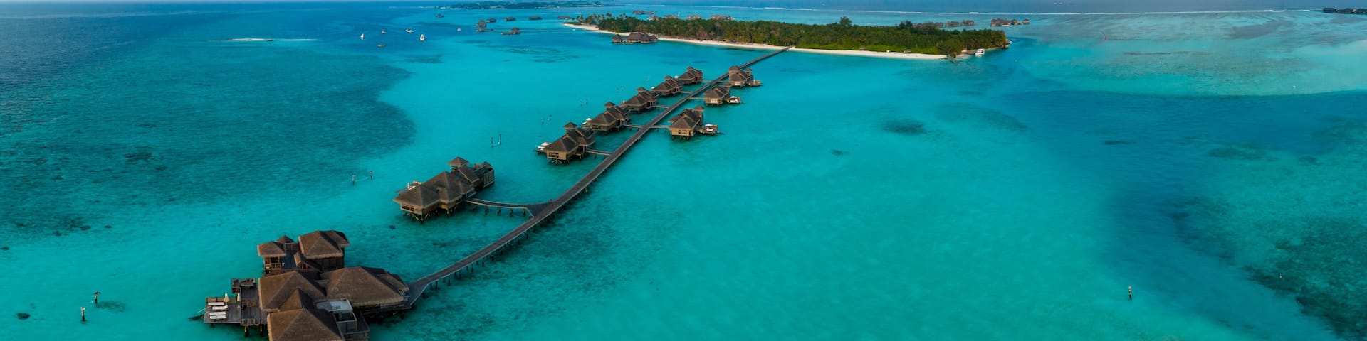 An aerial view of a tropical island resort with overwater bungalows connected by a wooden walkway.