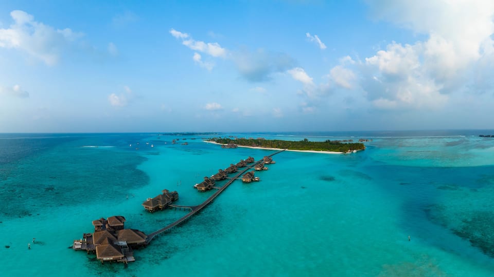 An aerial view of a tropical island resort with overwater bungalows connected by a wooden walkway.
