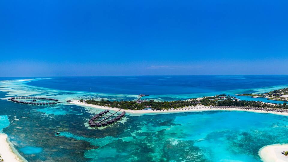 An aerial view of a tropical island with clear turquoise waters, overwater bungalows, and a white sandy beach.
