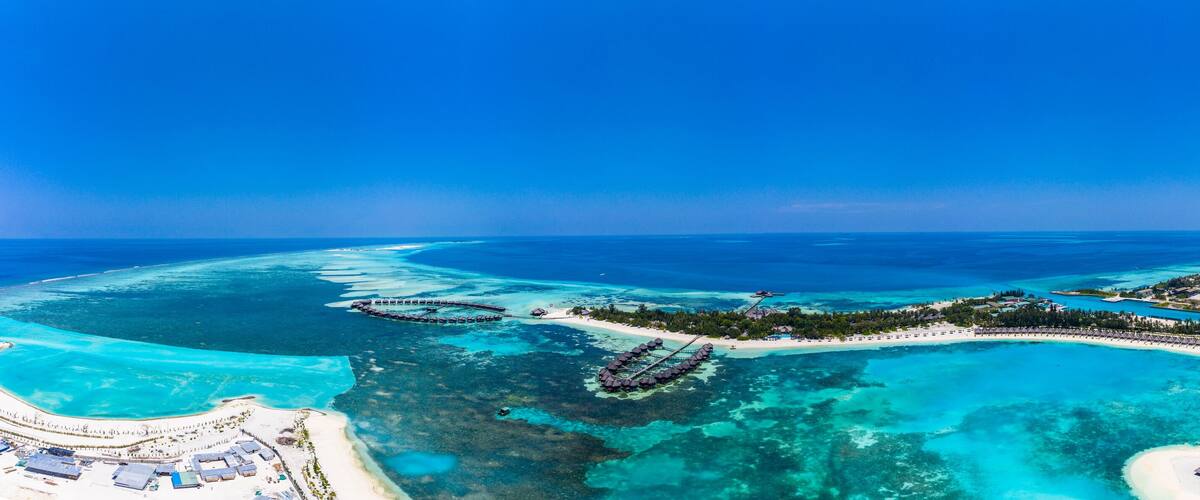An aerial view of a tropical island with clear turquoise waters, overwater bungalows, and a white sandy beach.