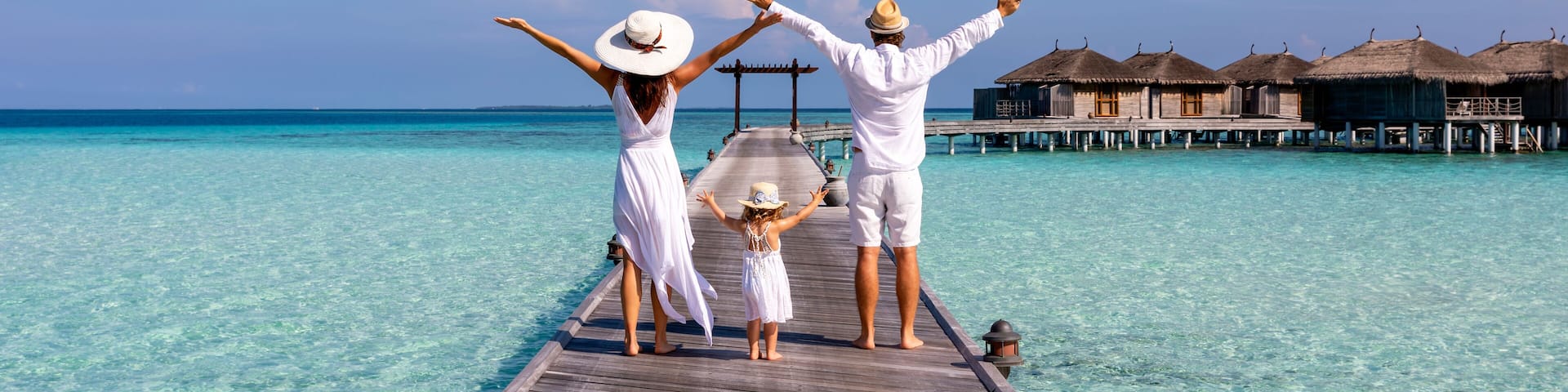 A happy family in white summer clothing on vacation walks along a wooden pier over tropical, turquoise ocean in the Maldives, Indian Ocean