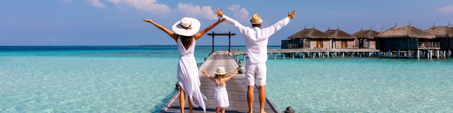 A happy family in white summer clothing on vacation walks along a wooden pier over tropical, turquoise ocean in the Maldives, Indian Ocean