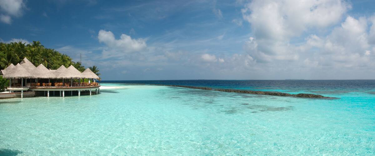 A beach with clear turquoise water, a thatched-roof structure, and palm trees.