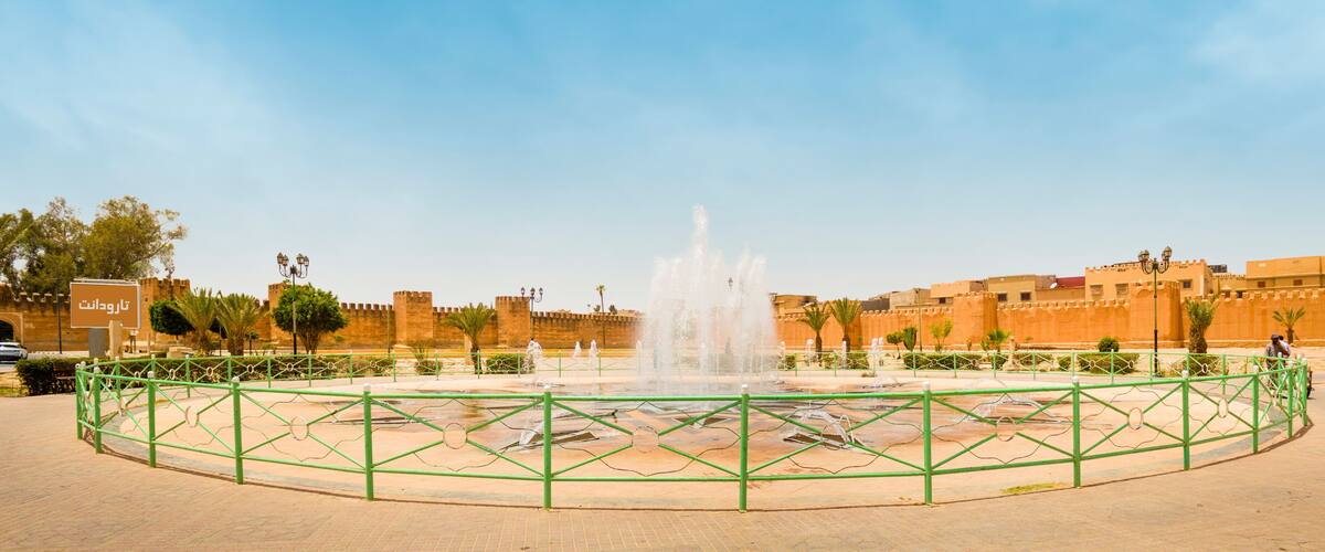 A historic fort with a central fountain, surrounded by a green fence and palm trees.
