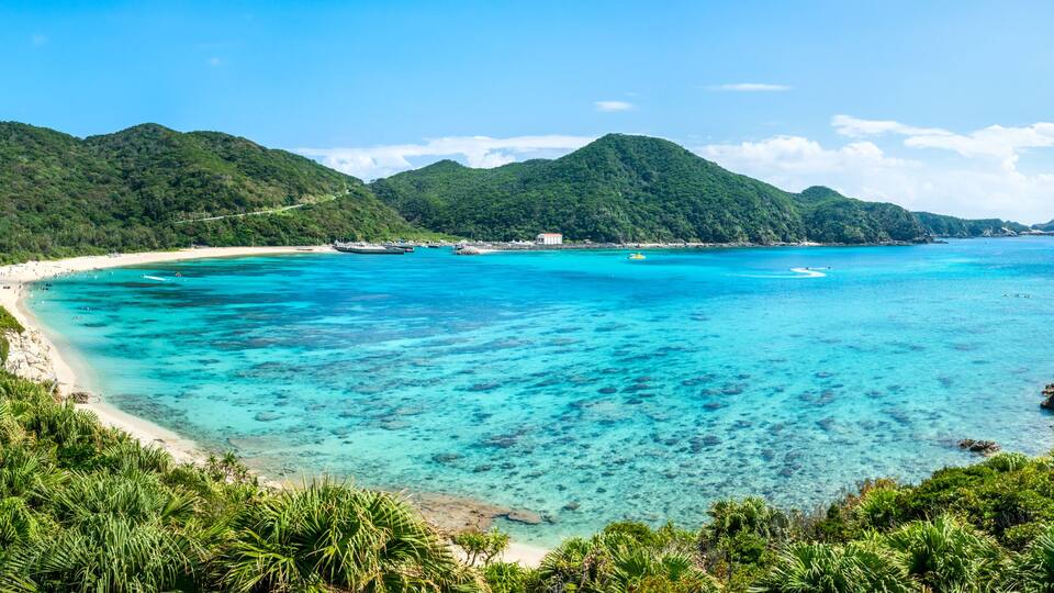 A tropical beach with clear turquoise waters, a sandy shore, and lush green hills in the background.