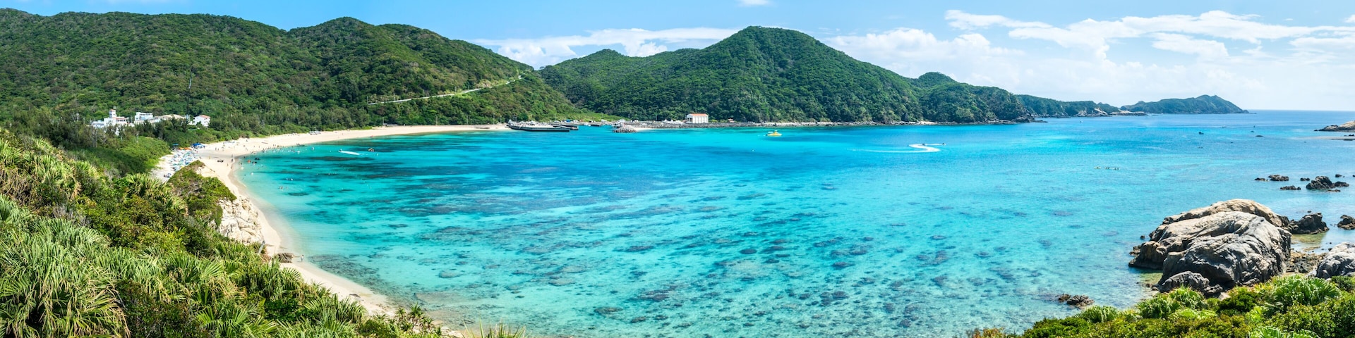 Ein tropischer Strand mit klarem türkisblauem Wasser, einem sandigen Ufer und üppigen grünen Hügeln im Hintergrund.