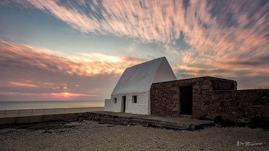 A white building with a stone structure, a beach, and a sunset.