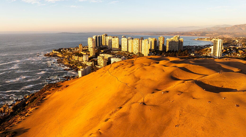 An aerial shot of Concón's sun-drenched dunes and coastal city, with people visible on the sandy landscape.