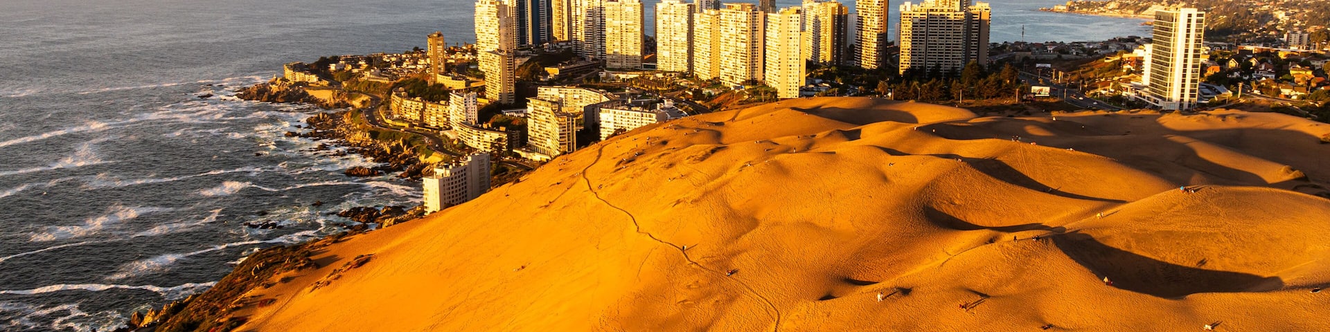 An aerial shot of Concón's sun-drenched dunes and coastal city, with people visible on the sandy landscape.