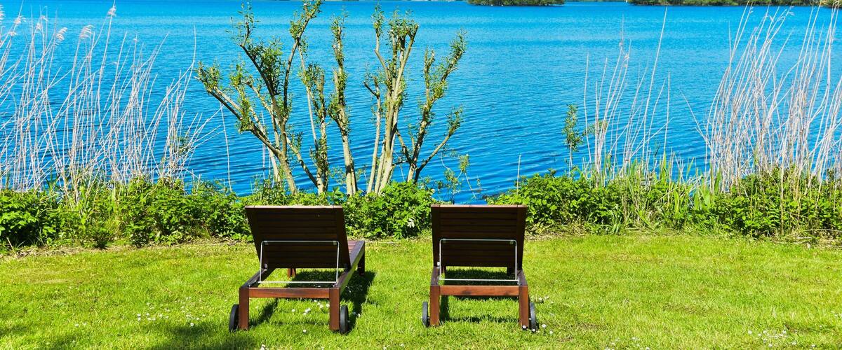 Two wooden chairs facing a large body of water with a clear sky and distant trees.