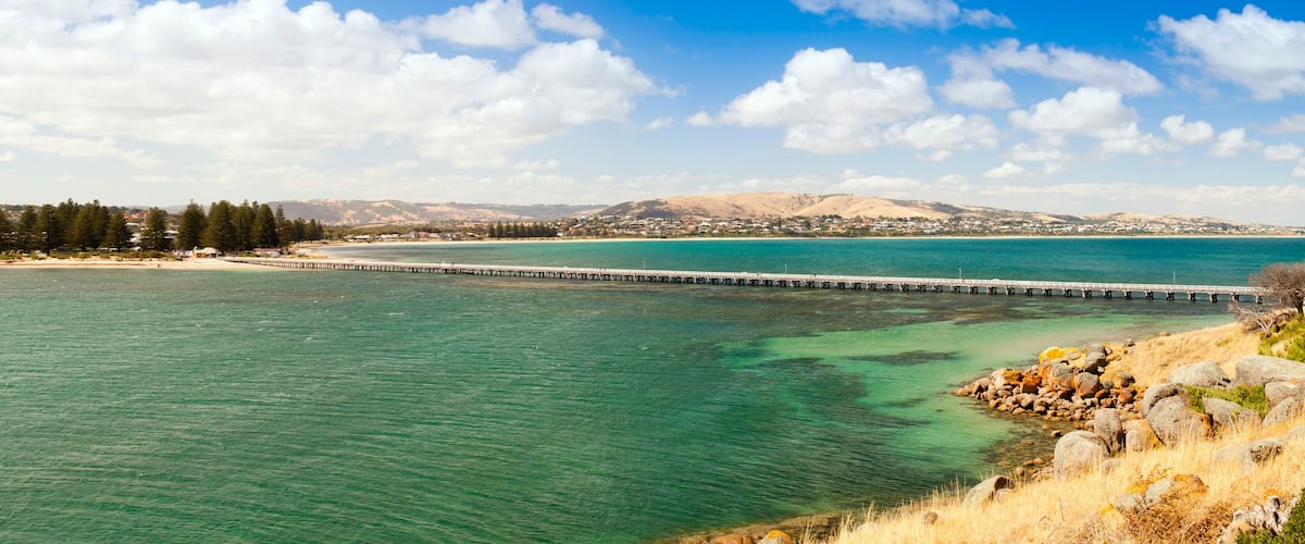 A coastal landscape with a pier extending into turquoise waters, a sandy beach, and a town in the distance.