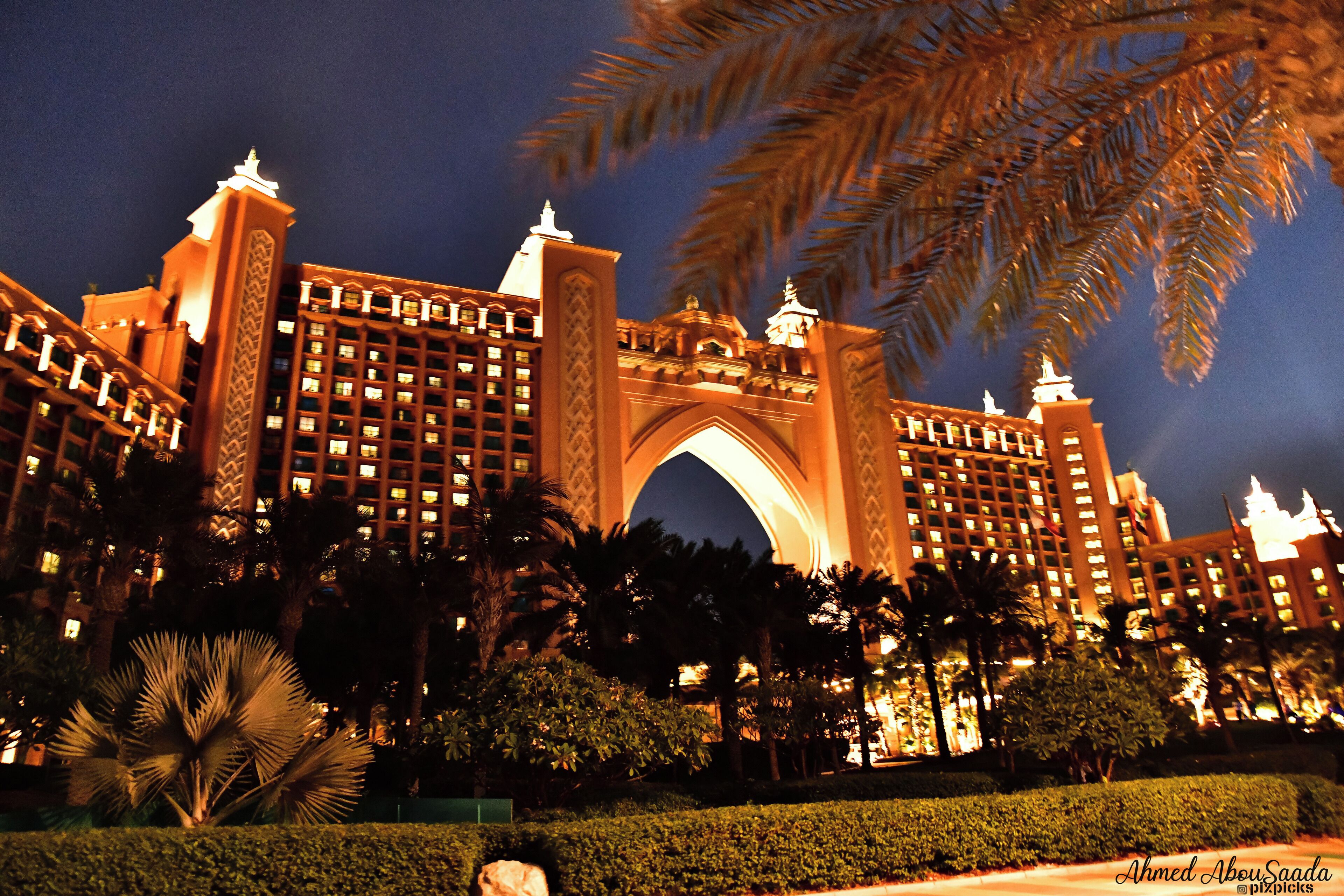 A grand hotel with an ornate archway, palm trees, and well-lit buildings at night.
