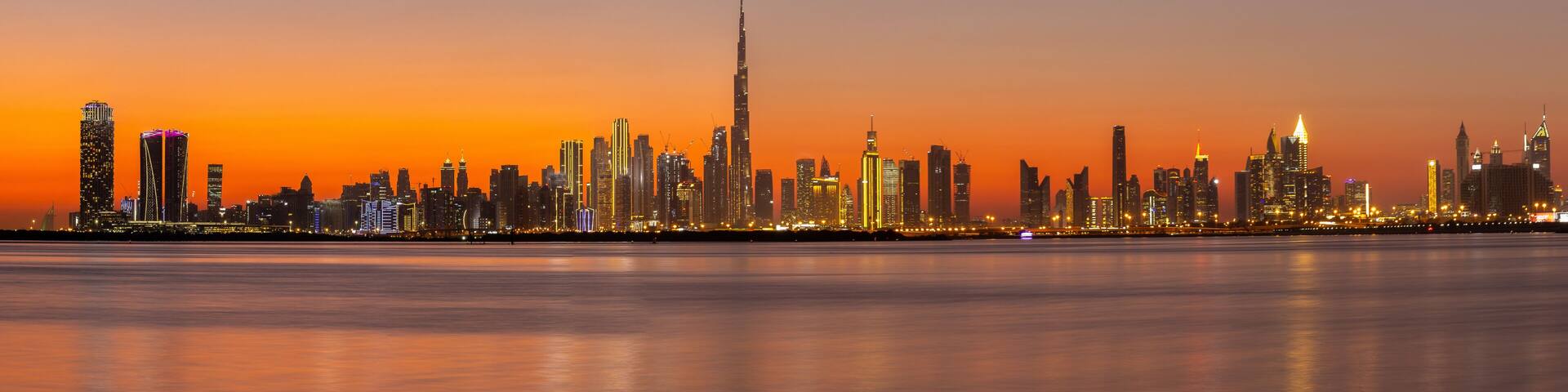 A city skyline at dusk with a prominent skyscraper in the center, a calm body of water in the foreground, and a gradient sky.
