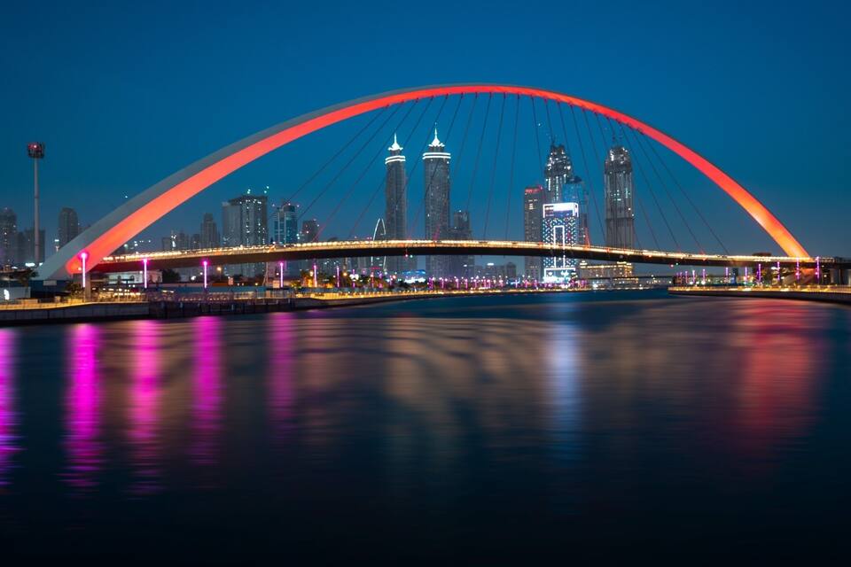 A brightly lit bridge with a red arch spanning over a body of water, with a city skyline in the background.