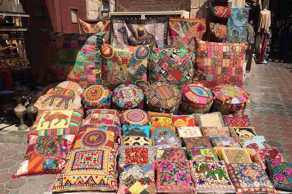 Una exhibición de almohadas y cojines de colores y diseños intrincados frente a un edificio con un letrero que dice "Bai Al Wakeel".