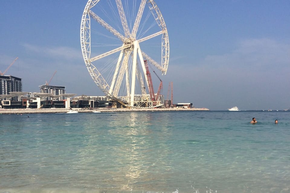 Uma praia com uma grande roda-gigante e guindastes de construção ao fundo.
